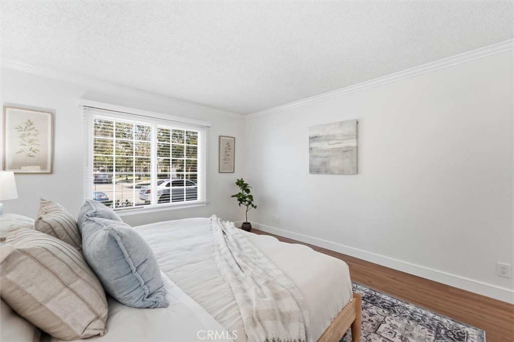Bright bedroom with white bedding, a large window with blinds, hardwood floors, and framed artwork.