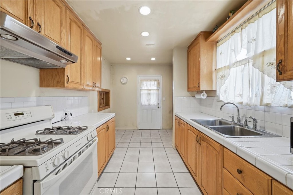 Narrow kitchen with honey-colored cabinets, white gas range, and double sink beneath a curtained window.