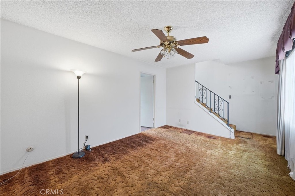 Empty living room with brown carpet, a ceiling fan, and a staircase with black metal railing.
