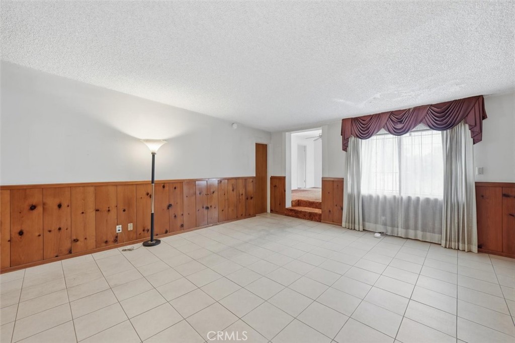 Empty living room with tiled floors, wood-paneled walls, and a large curtained window.