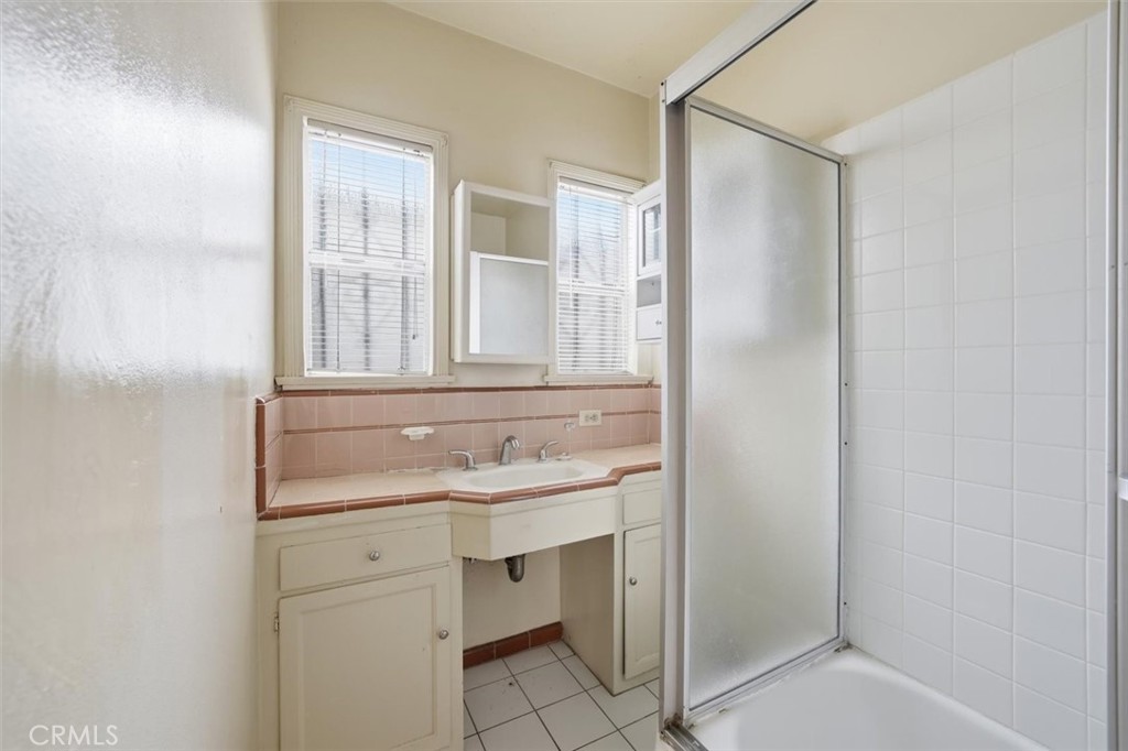 Bathroom with a cream vanity, pink tile countertop, two windows, and a frosted-glass shower door.