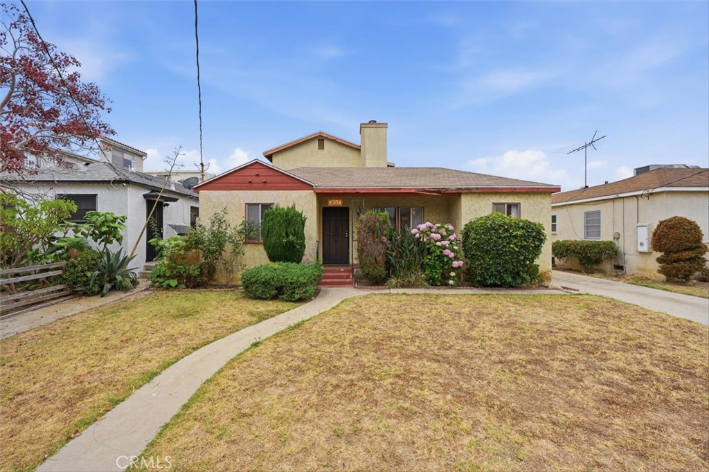 Single-story tan stucco house with red-trimmed gable, shrubs, and curved walkway across a dry front lawn.