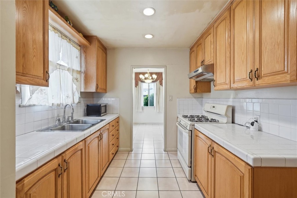 Galley kitchen with honey-colored wood cabinets, white tile counters, double sink, and gas range.