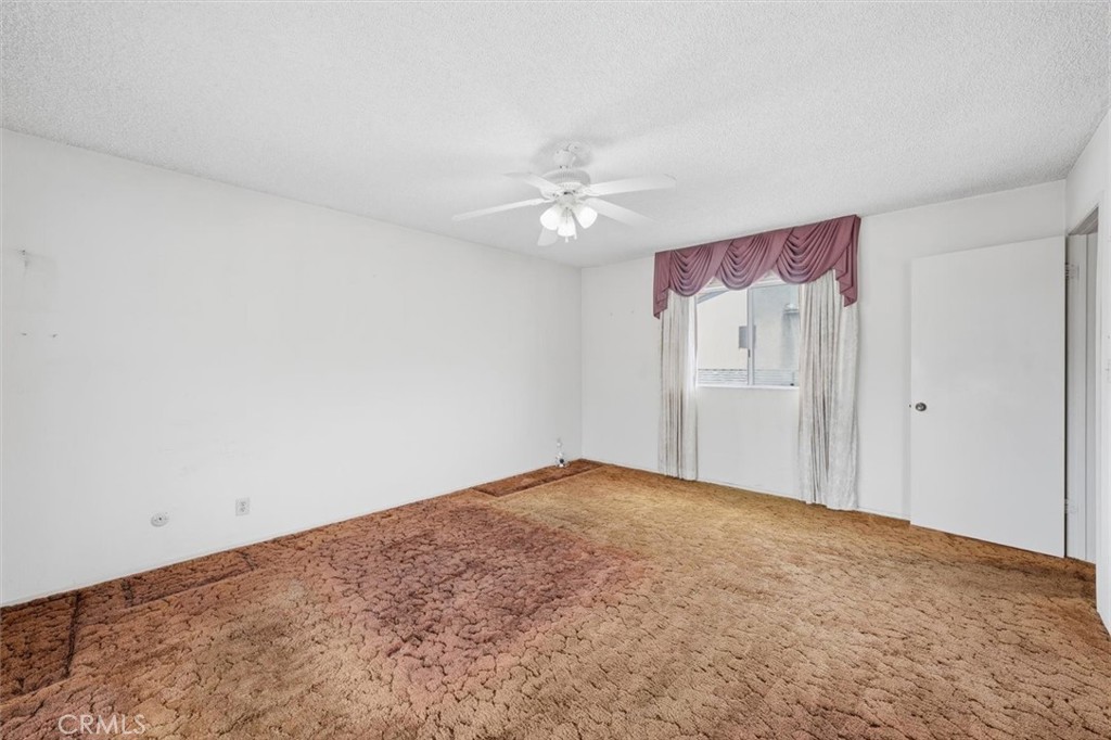 Empty bedroom with brown carpet, a ceiling fan, and mauve curtains over the window.