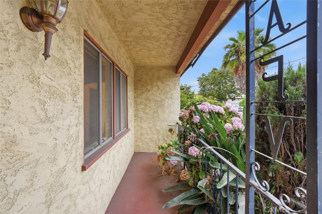Covered front porch with a stucco wall, red floor, and pink flowers beside a decorative metal railing.