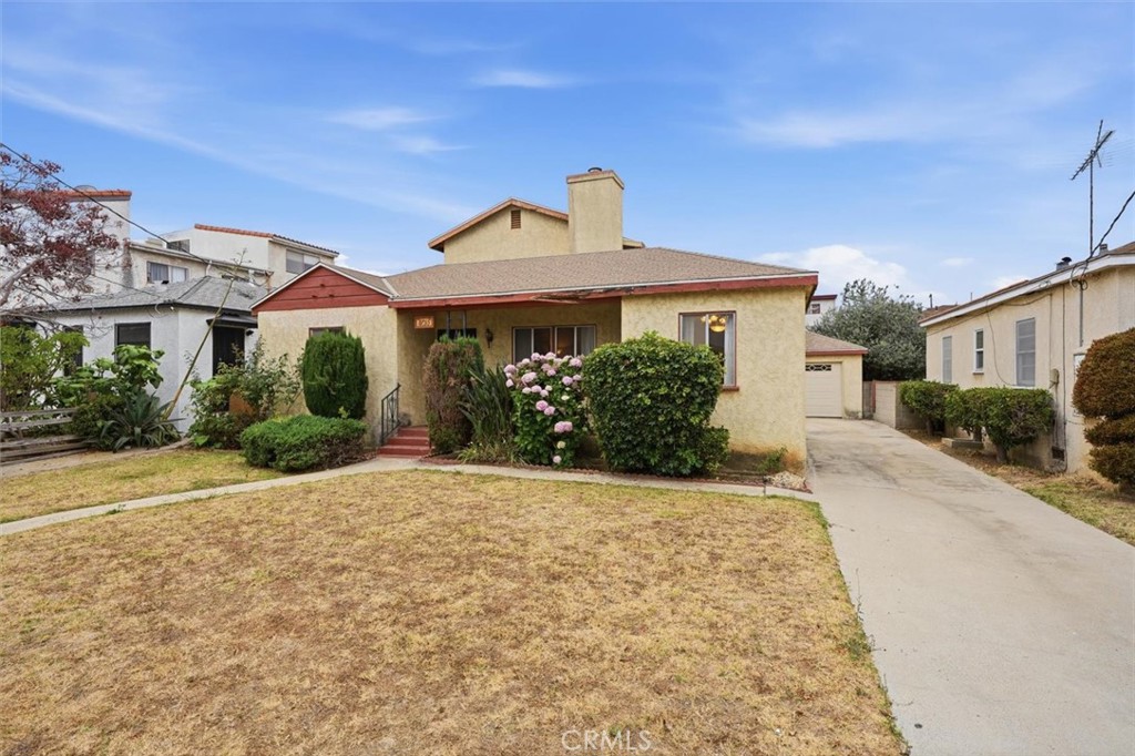 Single-story beige stucco house with a dry front lawn, flowering shrubs, and a driveway.