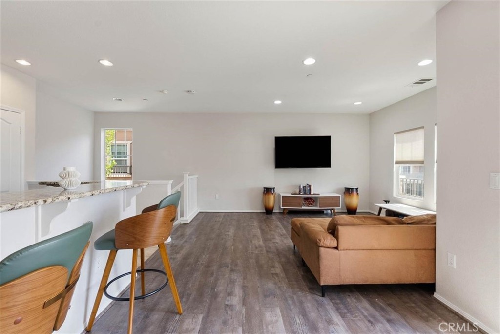 Open-plan living room with tan sectional, wall-mounted television, and breakfast bar with teal stools.