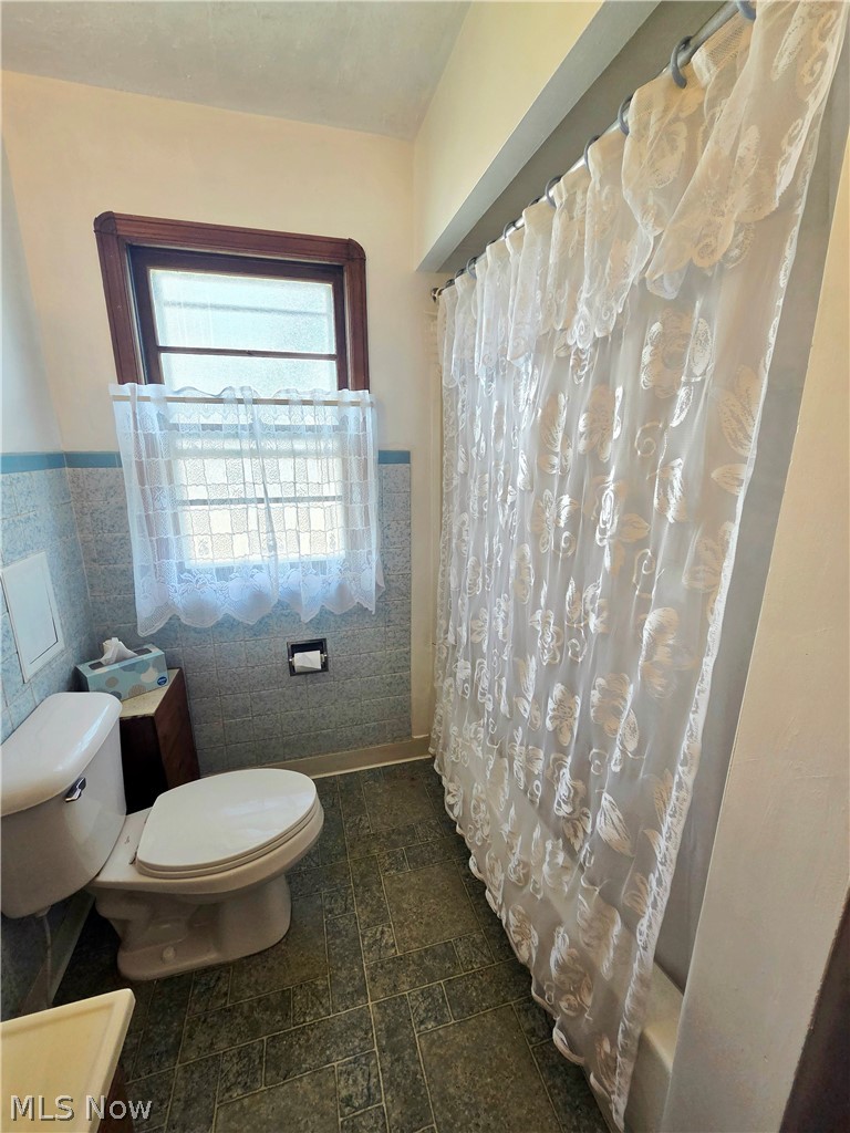 Bathroom with toilet, tiled walls and floor, and a patterned shower curtain beside a window.