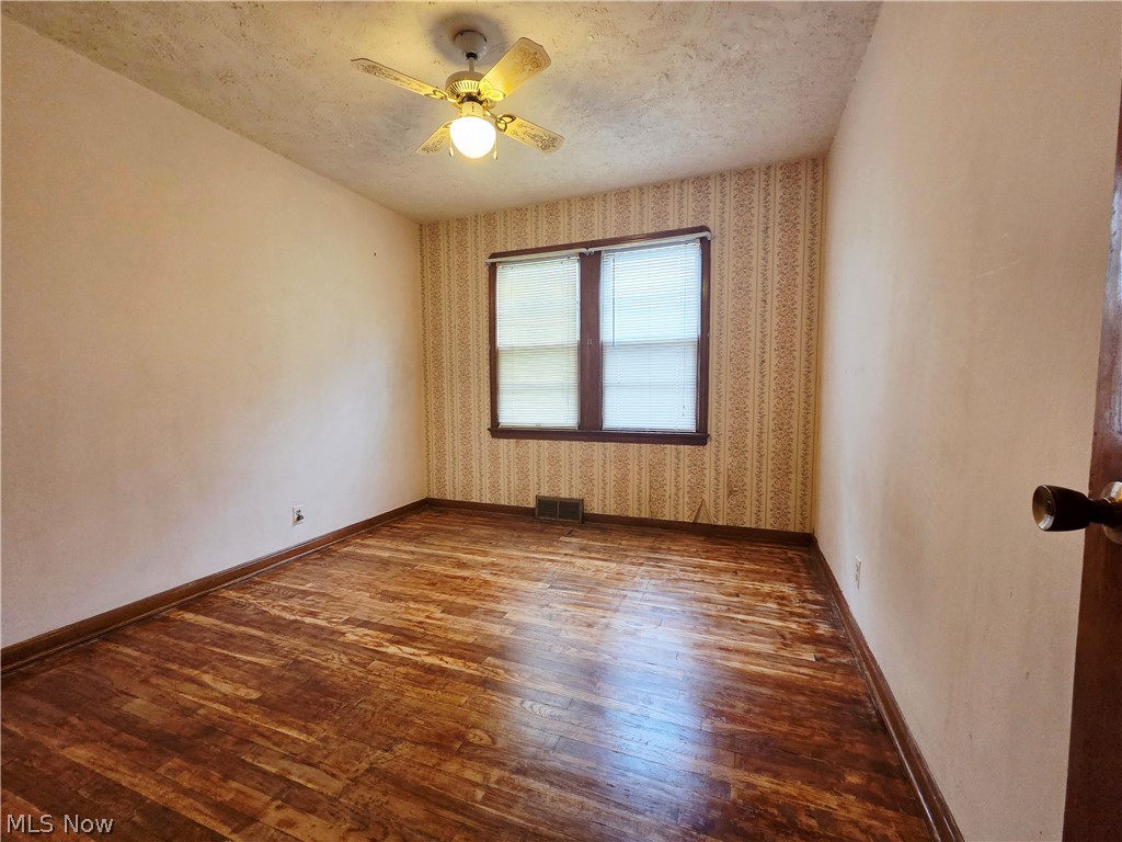 Empty bedroom with worn hardwood floors, patterned wallpaper, two windows, and a ceiling fan.