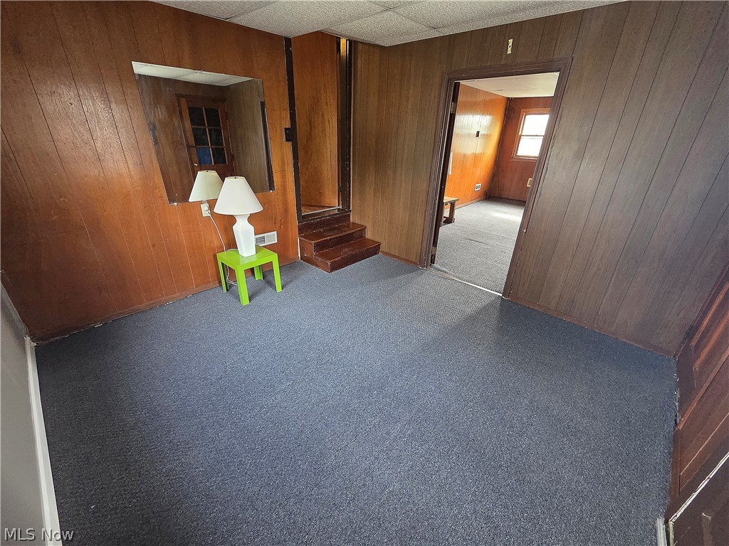 Wood-paneled room with gray carpet, a green table and lamp, and steps beside an open doorway.