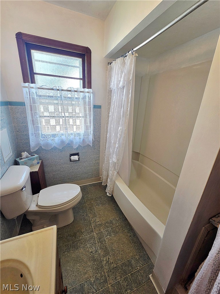 Bathroom with a lace-curtained tub-shower, toilet, and dark tiled floor.