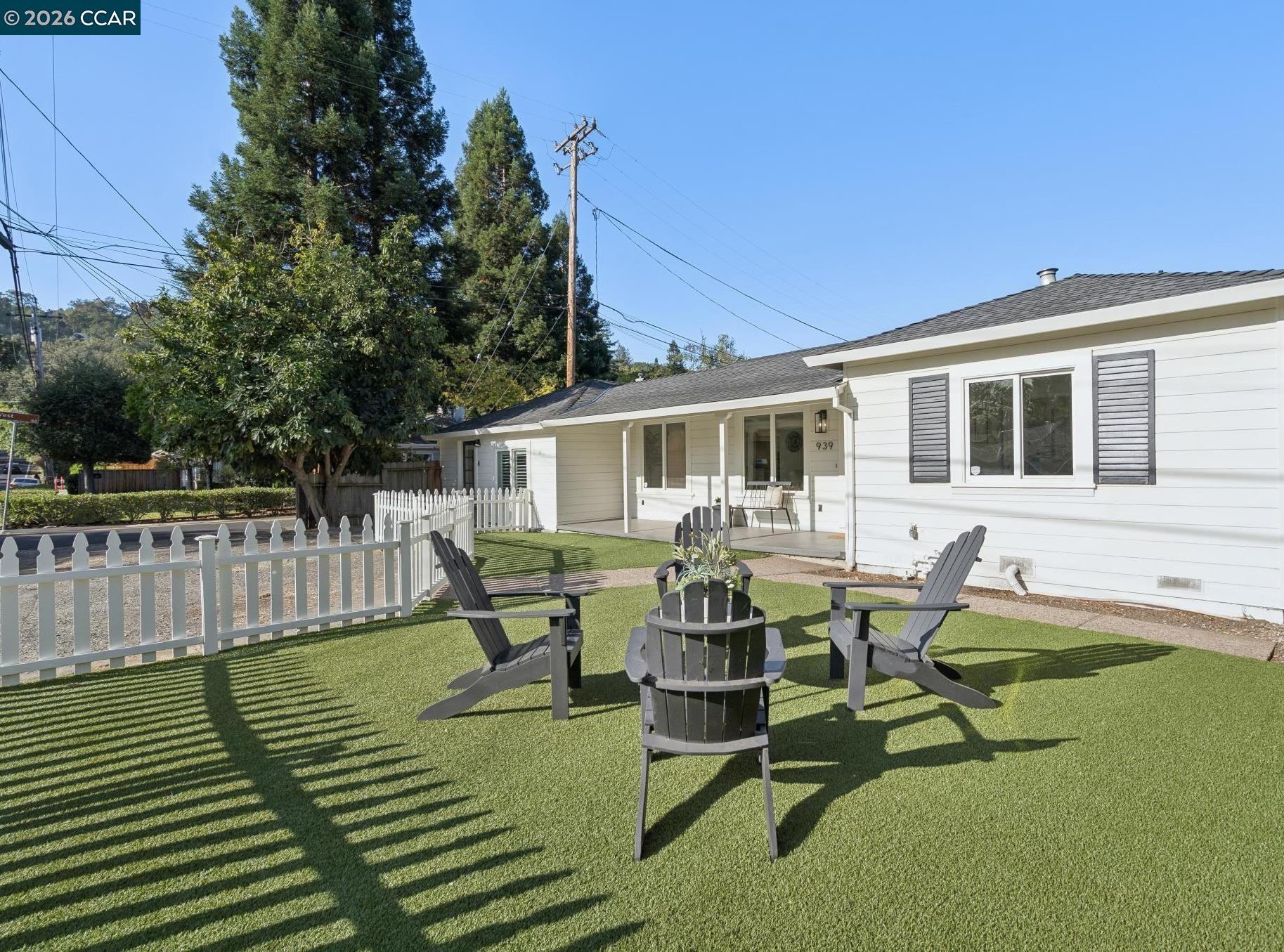 White single-story home with a covered porch, picket fence, and turf yard with outdoor seating.