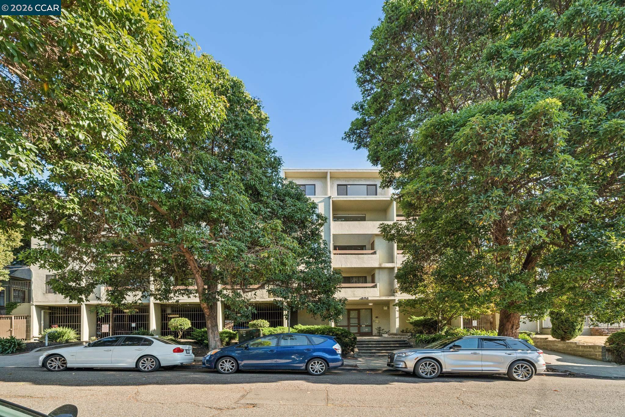 Multi-story condominium building with balconies, framed by leafy trees and parked cars.