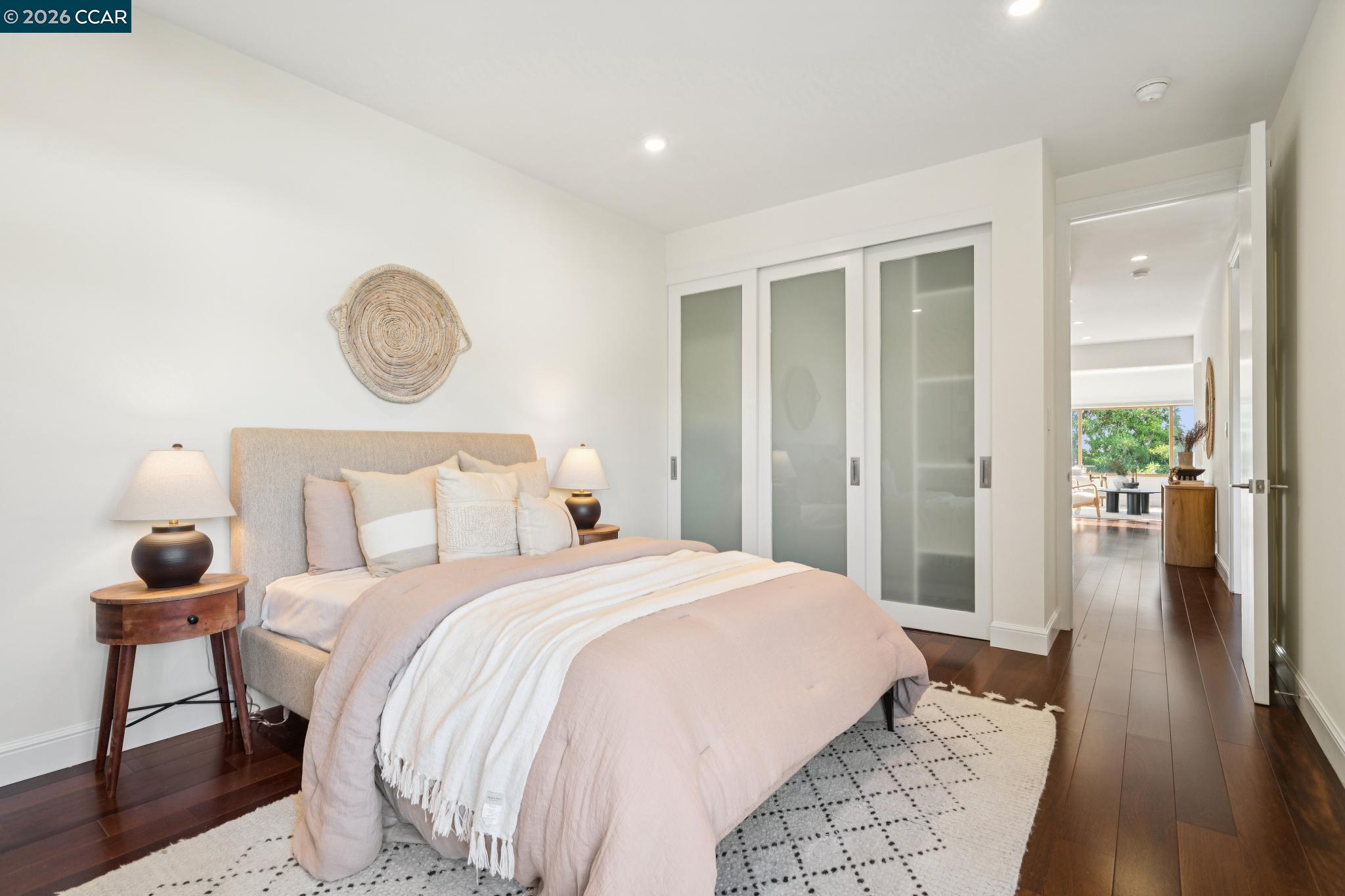 Staged bedroom with blush bedding, frosted-glass closet doors, and a view through the doorway to the living area.