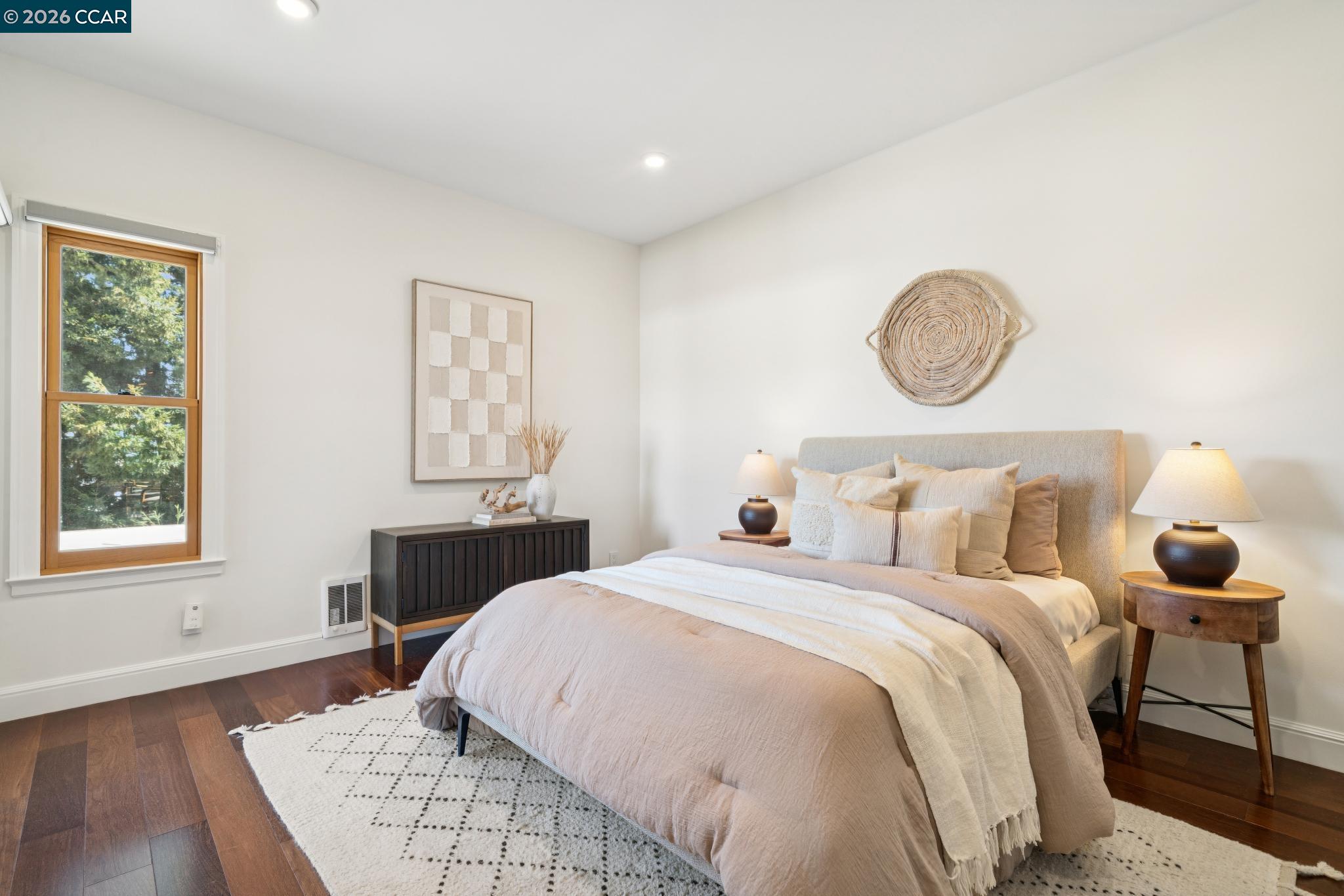 Staged bedroom with a neutral upholstered bed, bedside lamps, and a wood-framed window overlooking trees.
