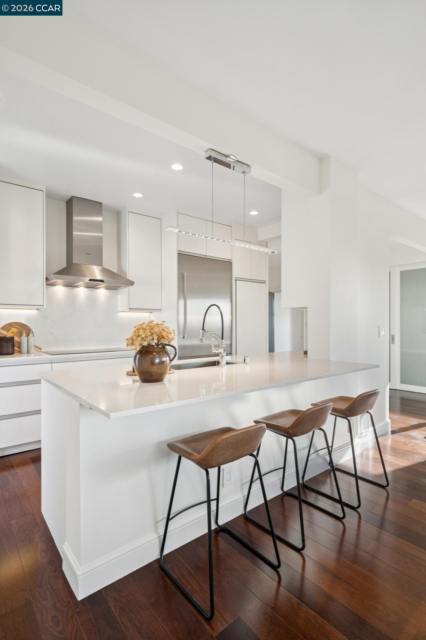 Modern white kitchen with a large island, three brown bar stools, stainless steel appliances, and dark hardwood floors.