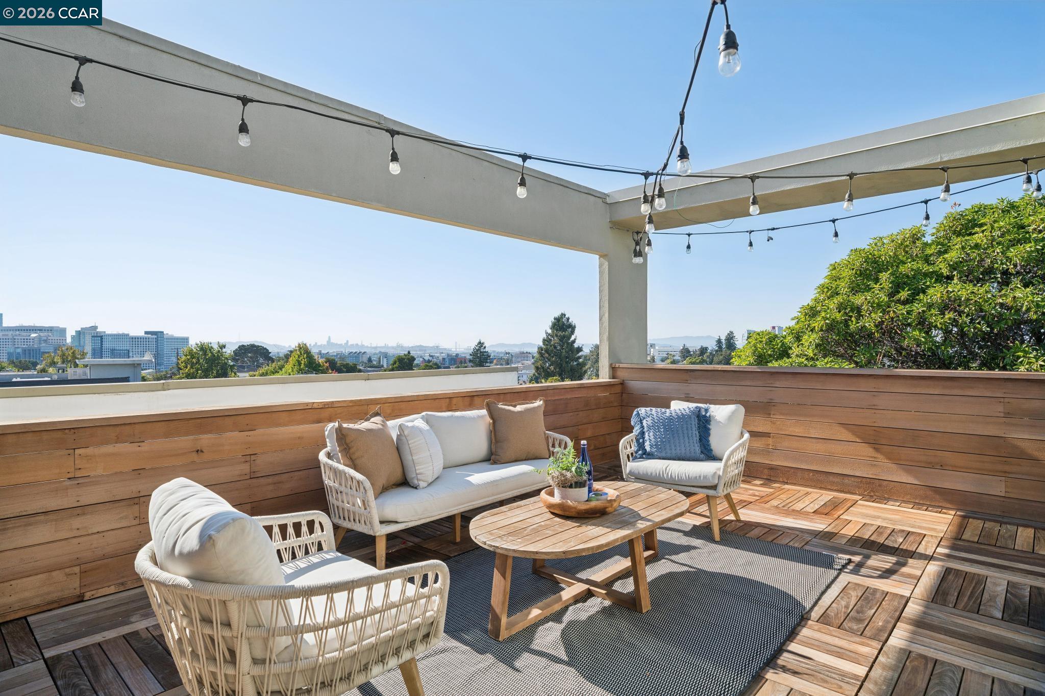 Rooftop deck with wicker seating, wood privacy walls, string lights, and distant city views.