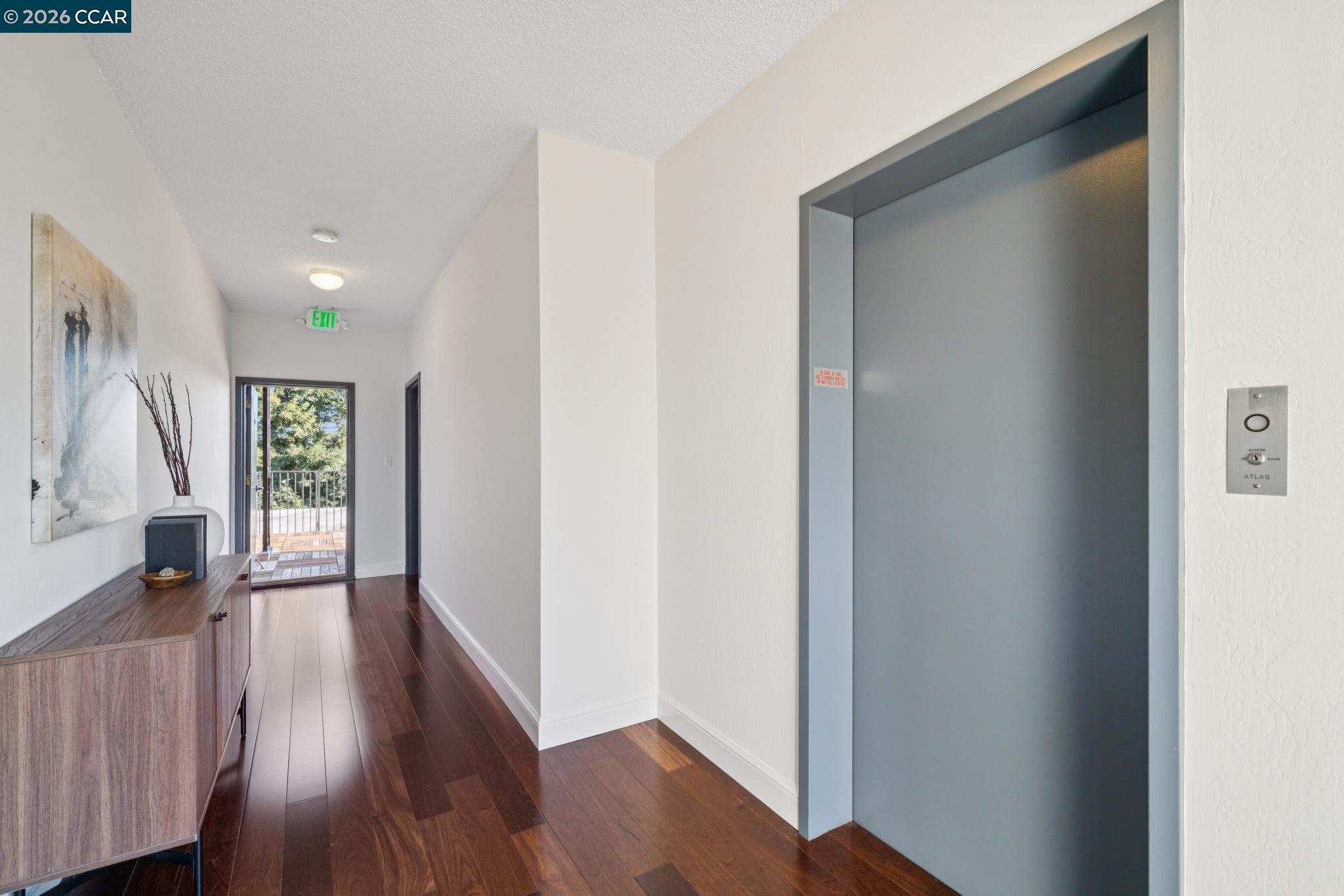 Condominium hallway with a gray elevator, dark wood floors, and a glass door to a balcony.