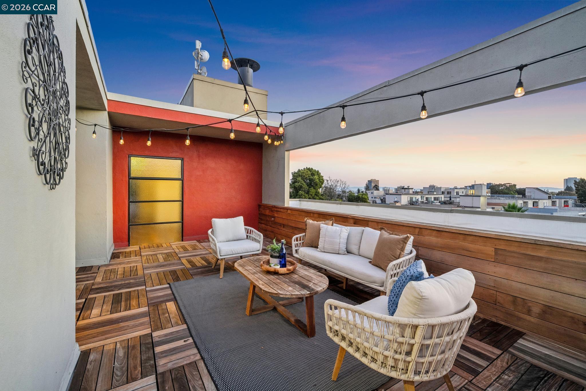 Rooftop terrace with woven lounge seating, wood decking, string lights, and city views at dusk.