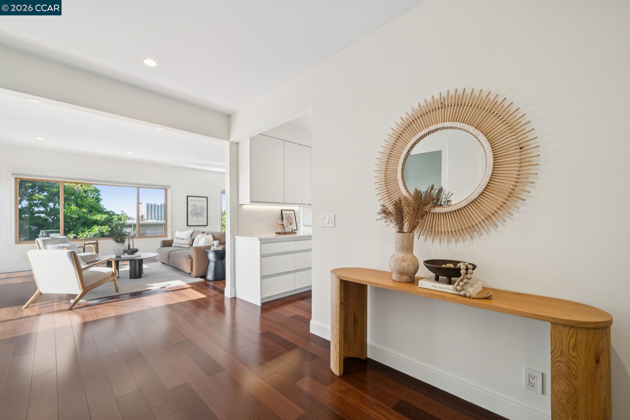 Staged condo living room and kitchen with dark wood floors, a large leafy-view window, and a sunburst mirror.