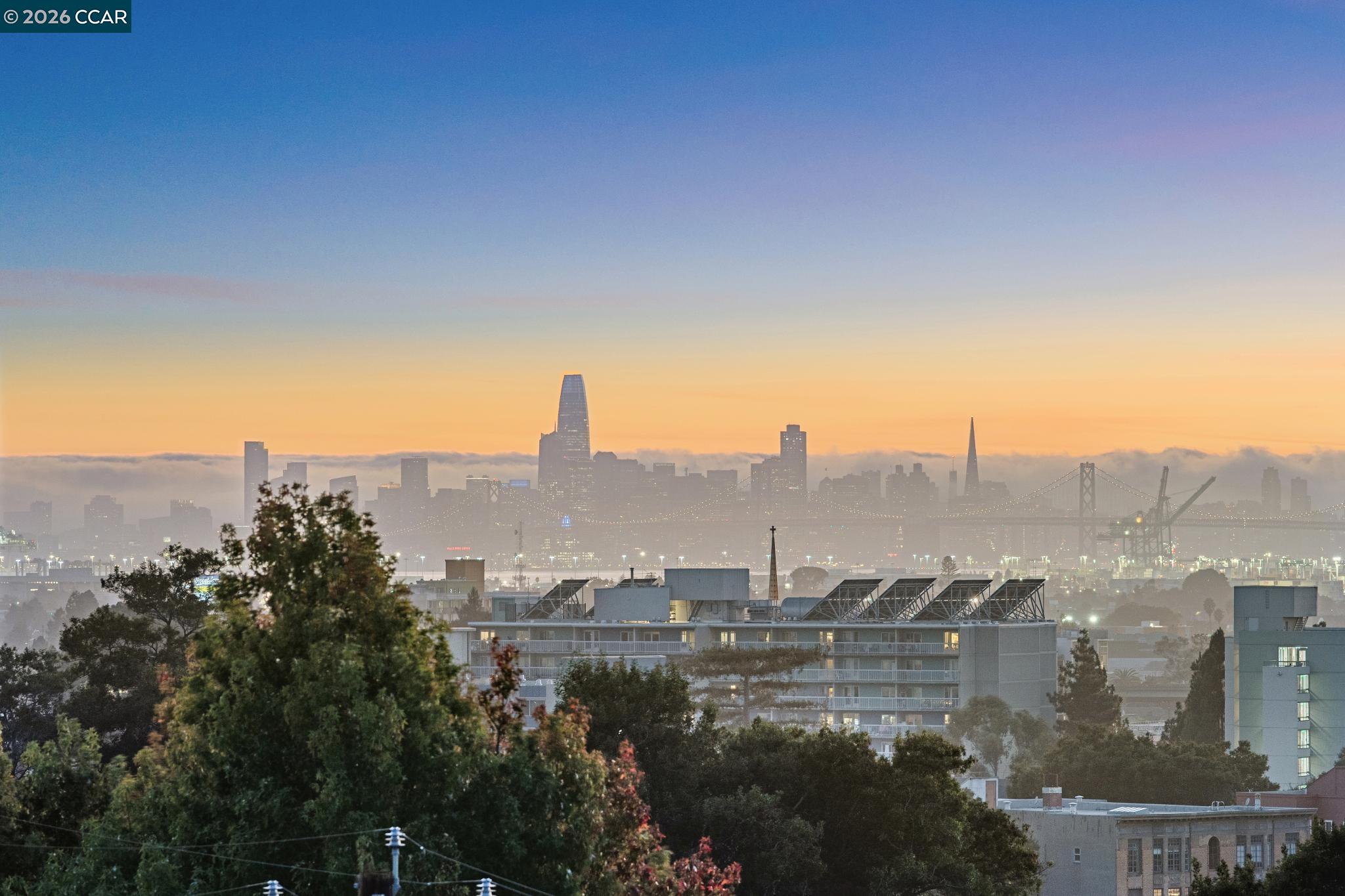 San Francisco skyline and Bay Bridge at sunset, seen above foreground rooftops and trees.