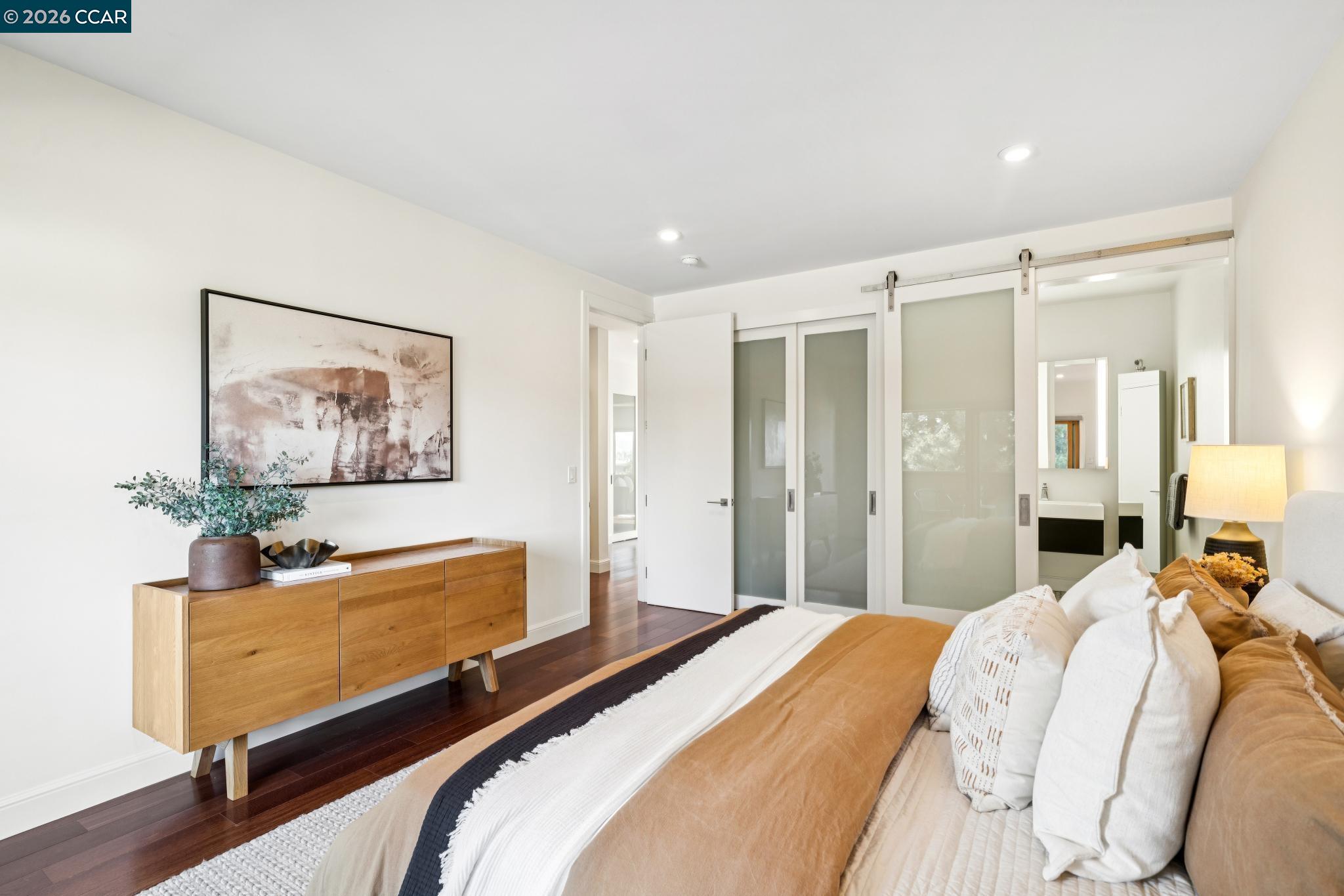 Staged bedroom with camel and white bedding, dark wood floors, and frosted-glass sliding doors.