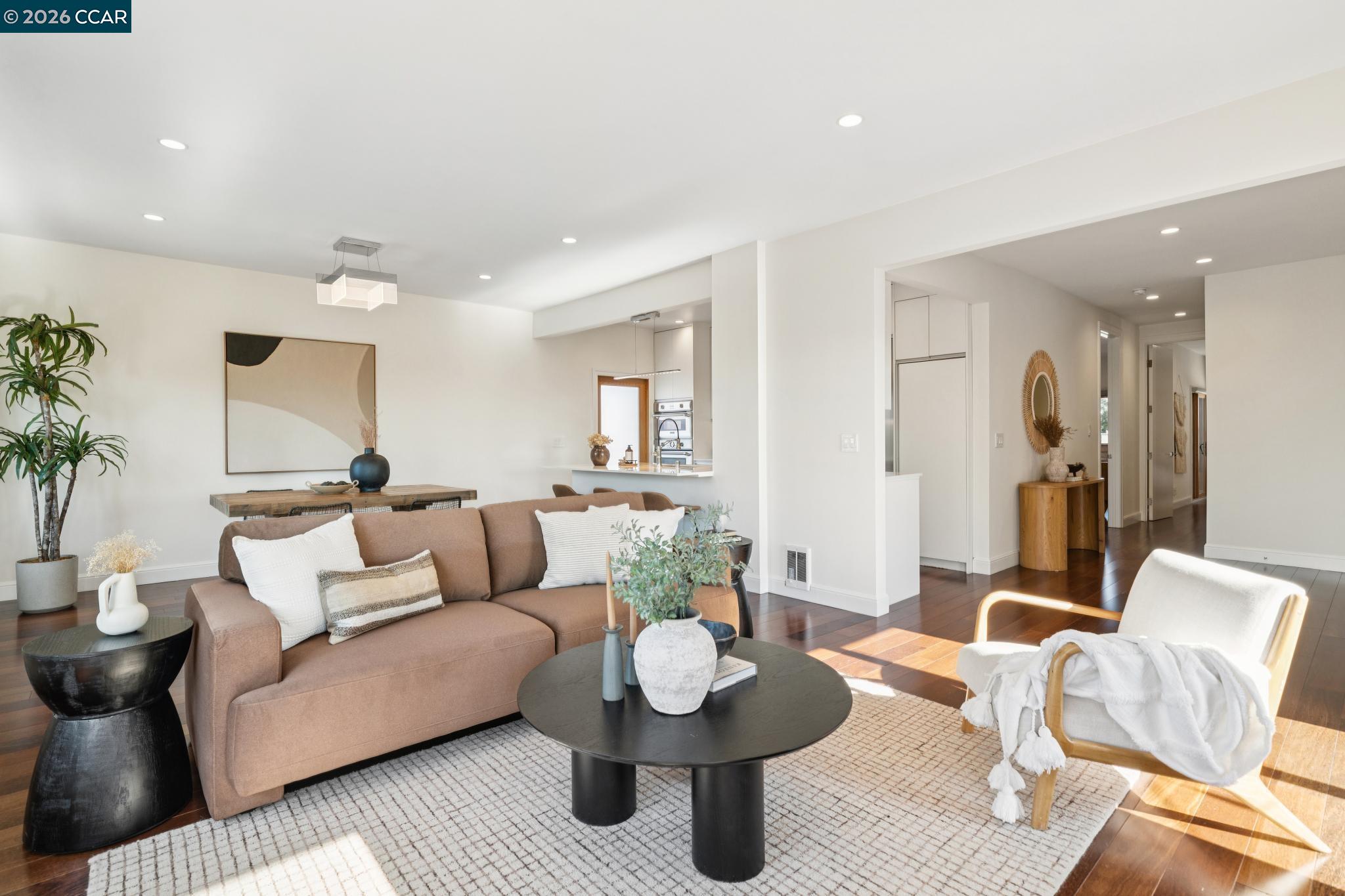 Furnished living room with a brown sofa, white accent chair, and sunlit hardwood floors.