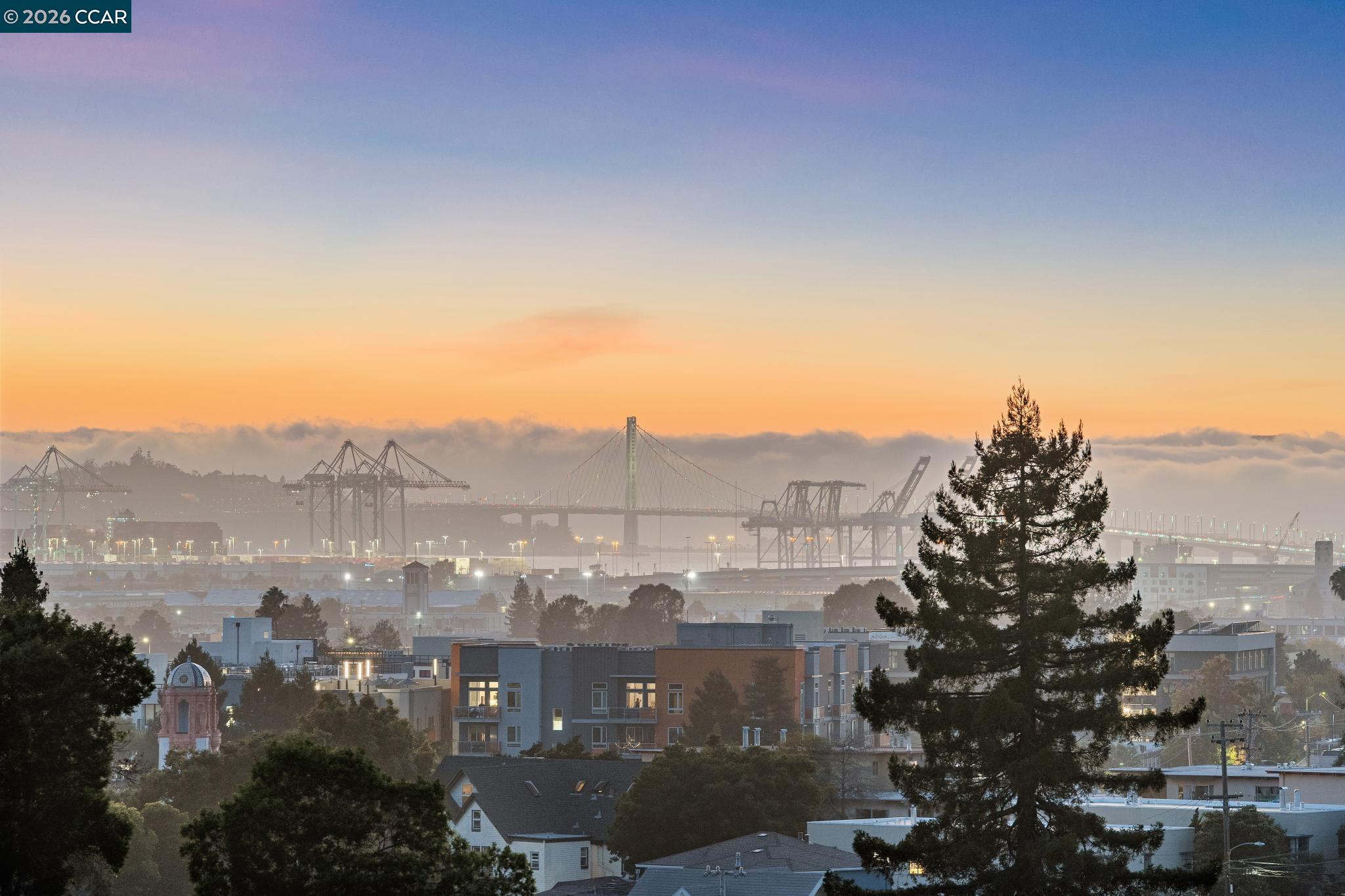 Oakland skyline and port cranes at sunset, with a suspension bridge above the fog.