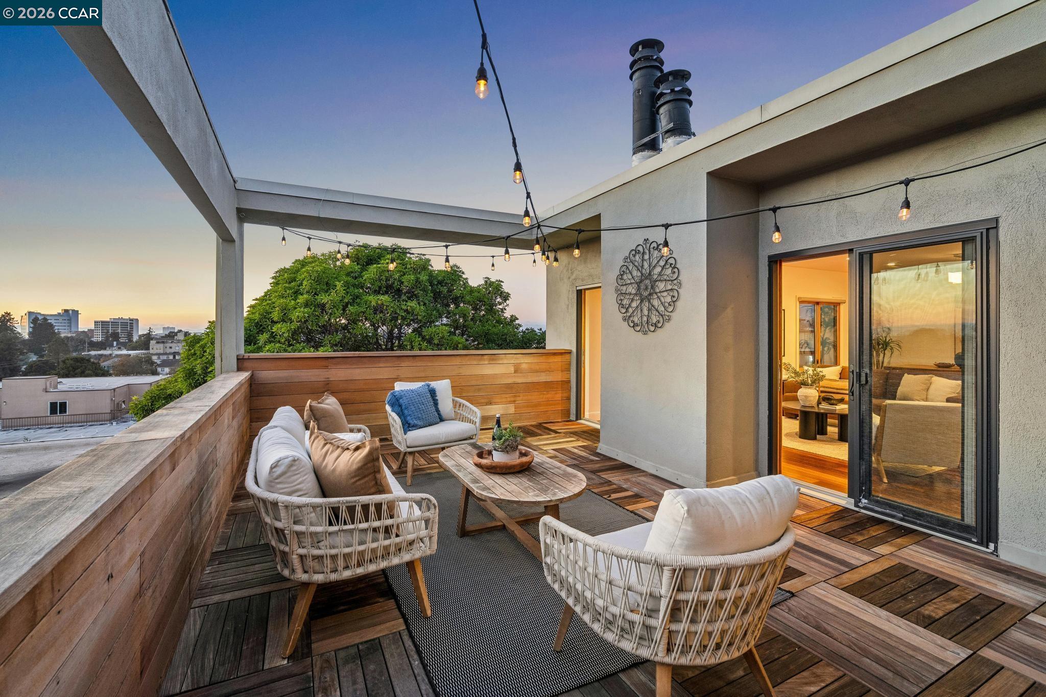 Rooftop deck with wicker seating, string lights, and an Oakland city view at dusk.