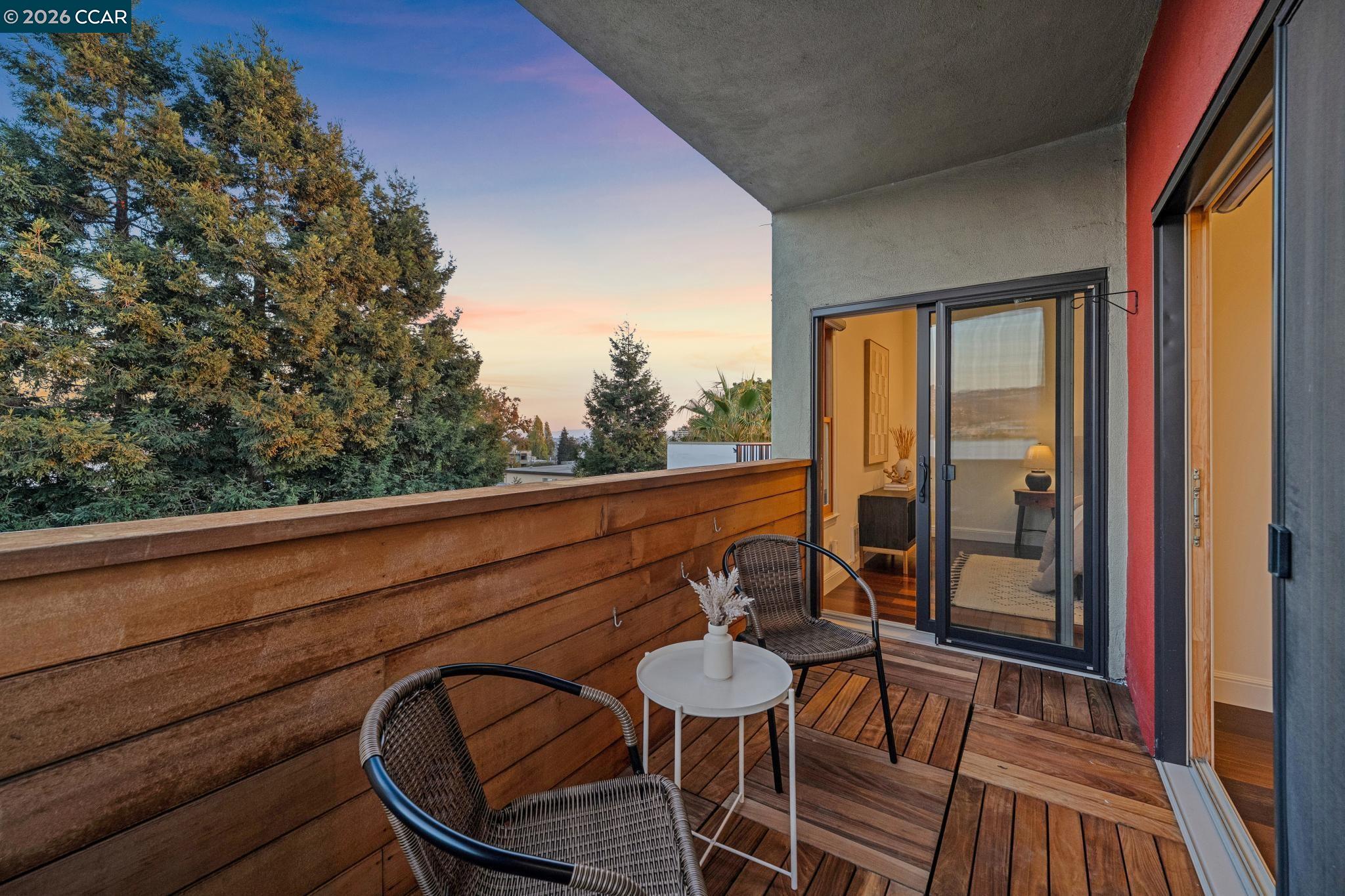 Apartment balcony with wicker seating and wood decking, opening to a view of trees at sunset.