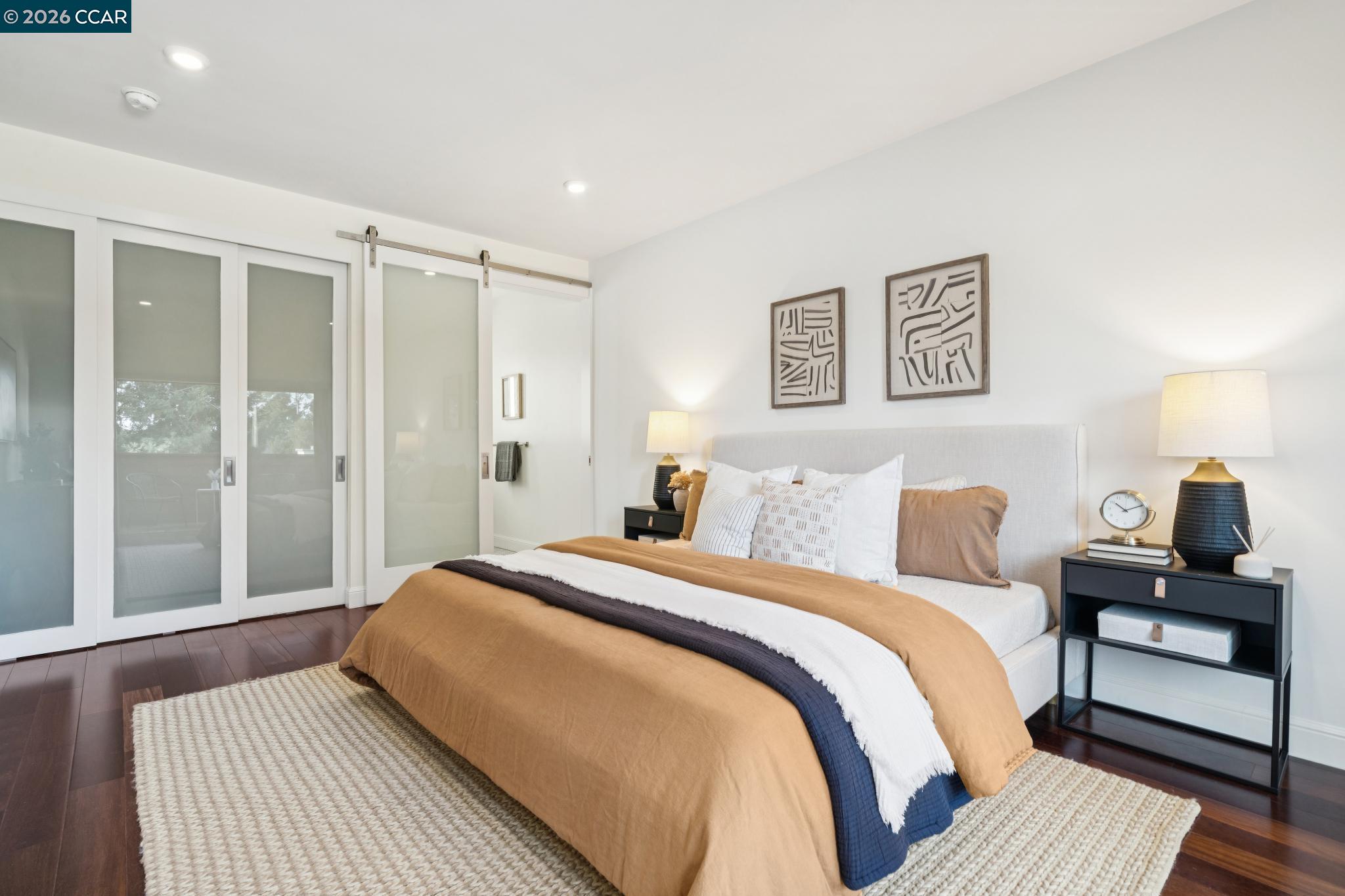 Staged bedroom with a tan-and-white bed, frosted-glass sliding doors, and dark wood floors.