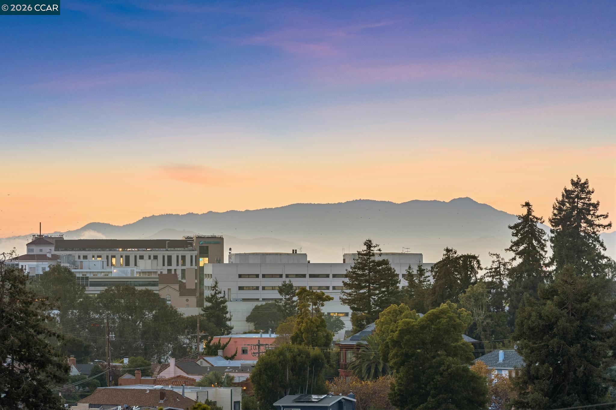 Oakland neighborhood rooftops and buildings with a mountain ridge in the distance at sunset.