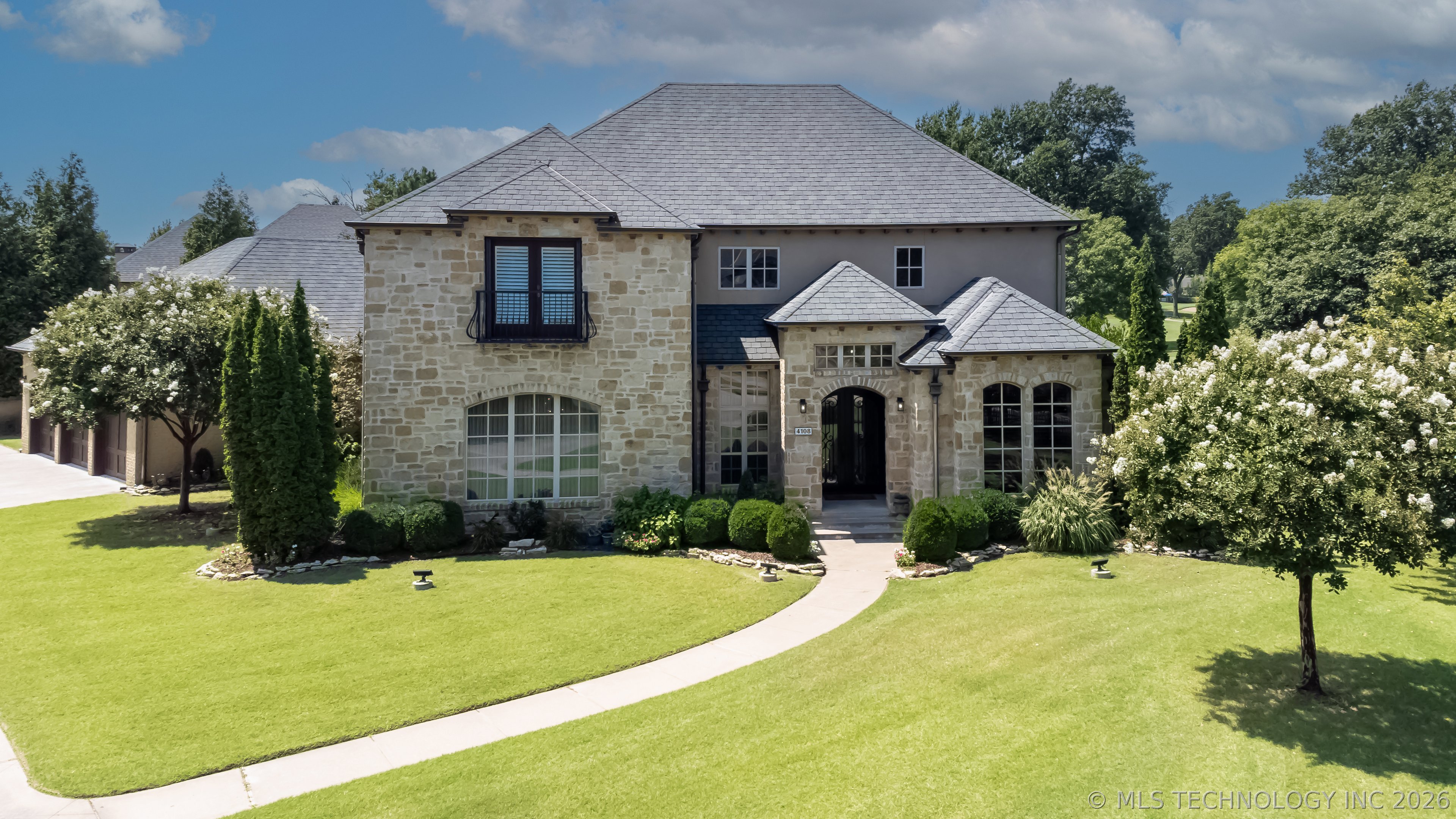 Two-story stone-and-stucco home with arched windows, landscaped lawn, and a curved front walkway.