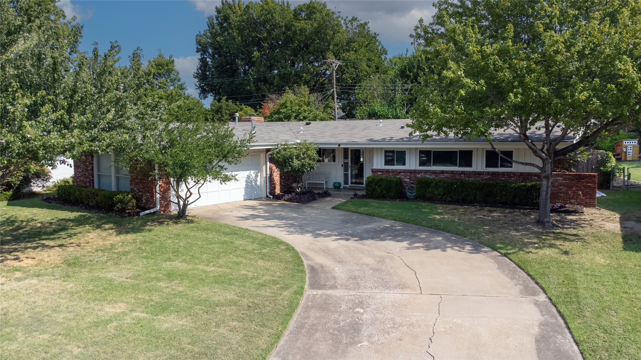 Single-story brick-and-white house with an attached garage, curved driveway, and mature trees.