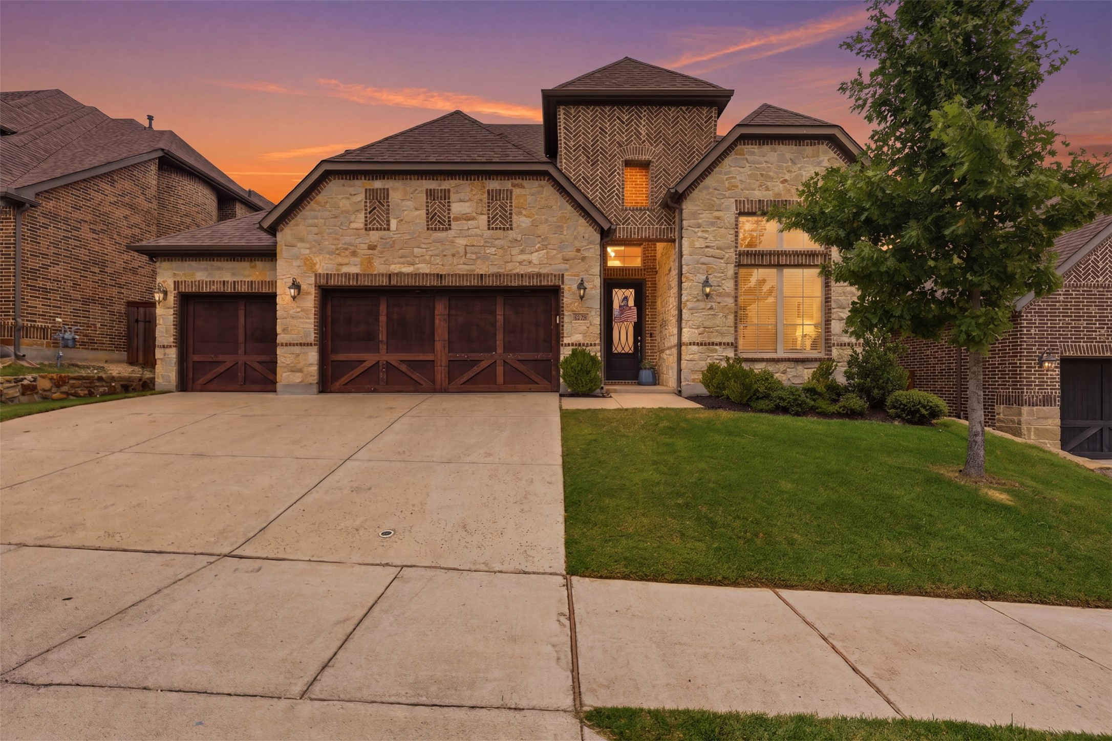 Two-story stone-and-brick home with three wood garage doors beneath an orange dusk sky.