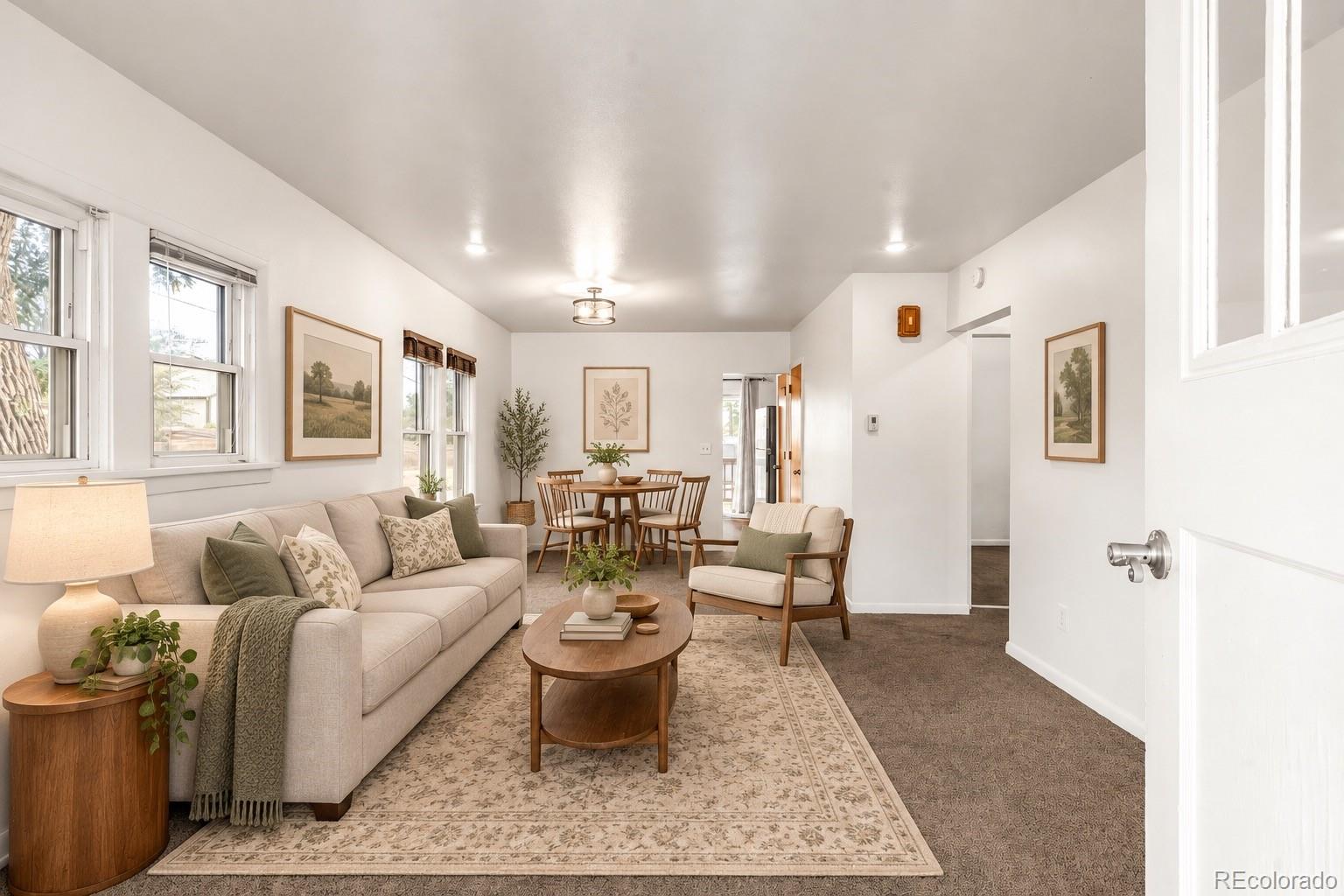 Staged living and dining room with a beige sofa, wood furniture, patterned rug, and bright windows.
