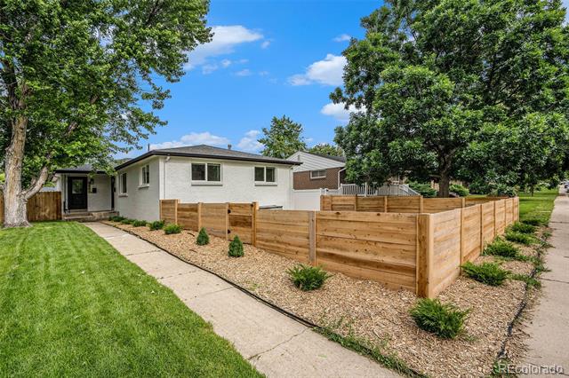 Single-story white house behind a wooden privacy fence, with a curved walkway and mature trees.