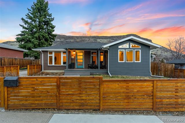 Single-story gray-and-brick house behind a wooden fence, with a mountain ridge at sunset.