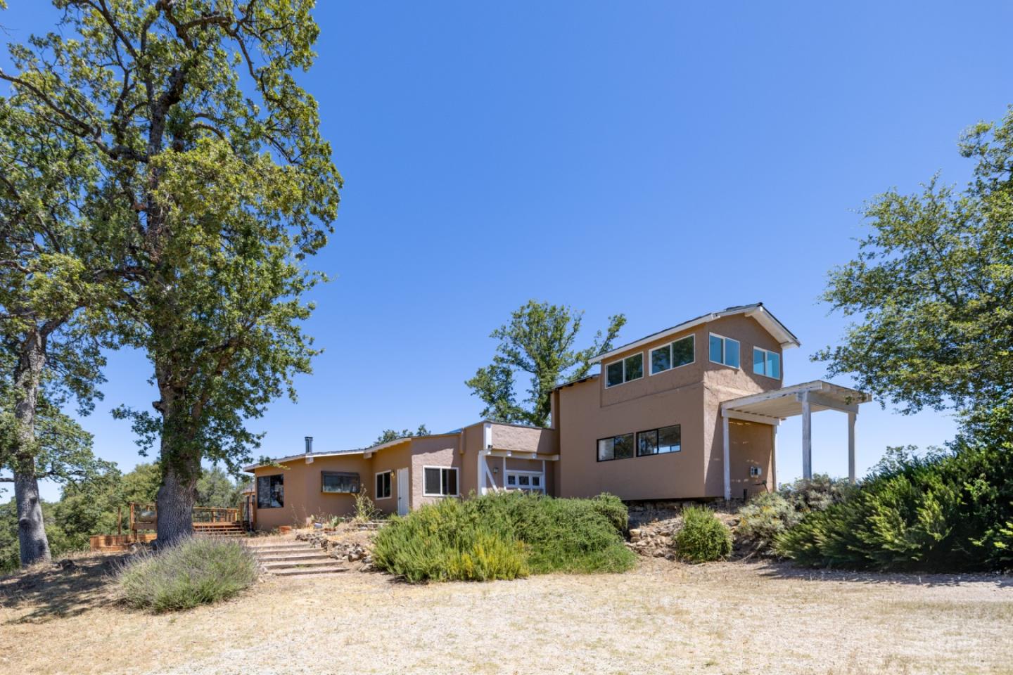 Two-story tan home with a covered entry, mature trees, and dry landscaped grounds.