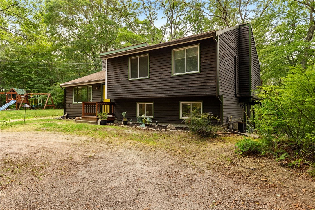 Dark brown two-story house in a wooded setting, with a gravel driveway and backyard playset.