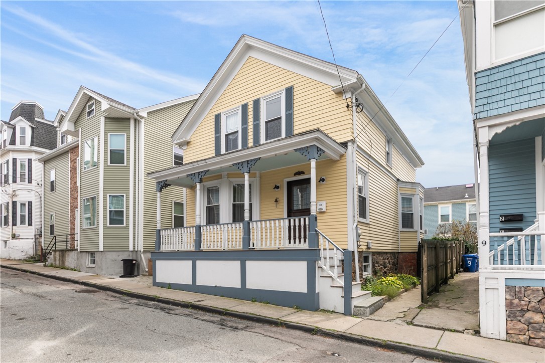 Yellow clapboard house with a covered porch and white railings on a narrow residential street.