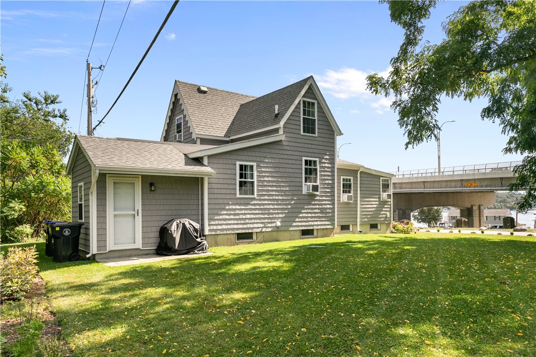 Gray shingle-sided house with white trim and gabled roof beside a lawn, with a bridge and water in the background.