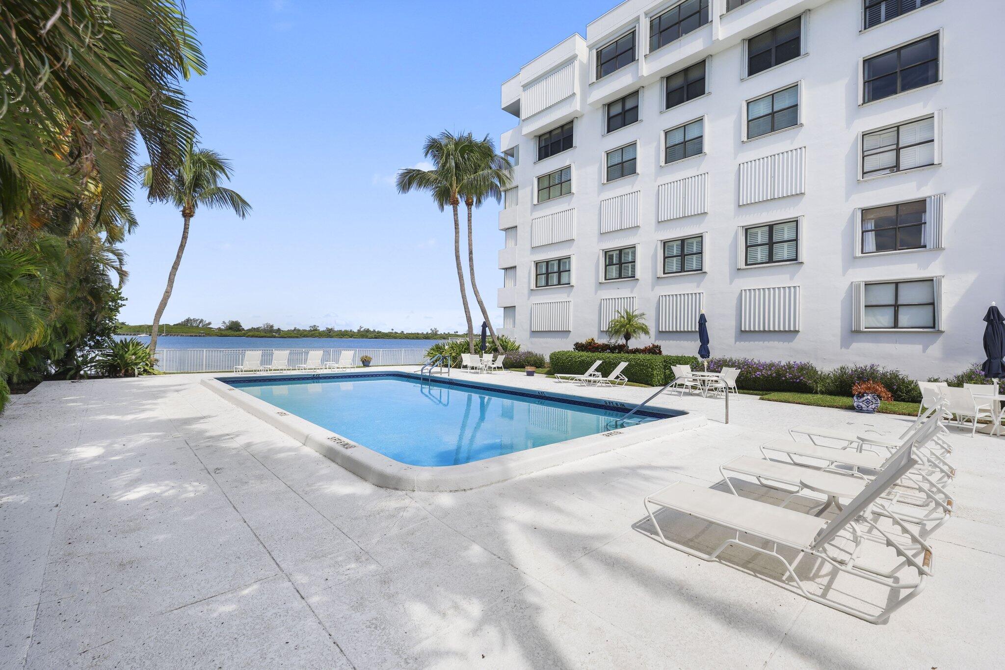 Condominium pool deck with palm trees, white lounge chairs, and waterfront views.