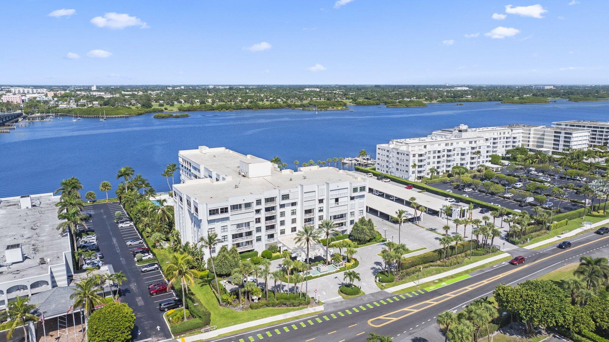 Waterfront condominium buildings beside a broad blue waterway, with palm-lined grounds and the far shore beyond.