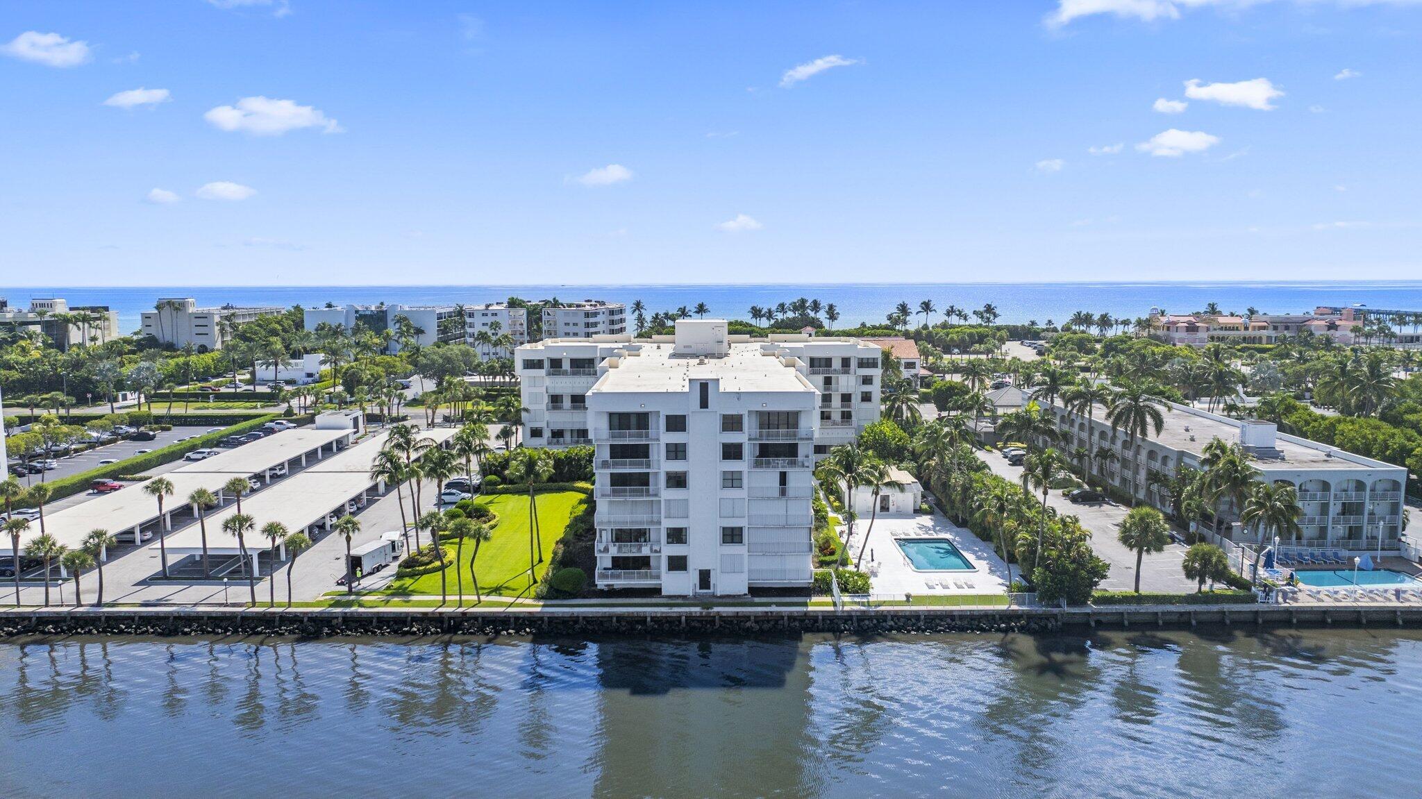 Aerial view of a white condominium between the ocean and a foreground waterway, with a pool and palm trees.