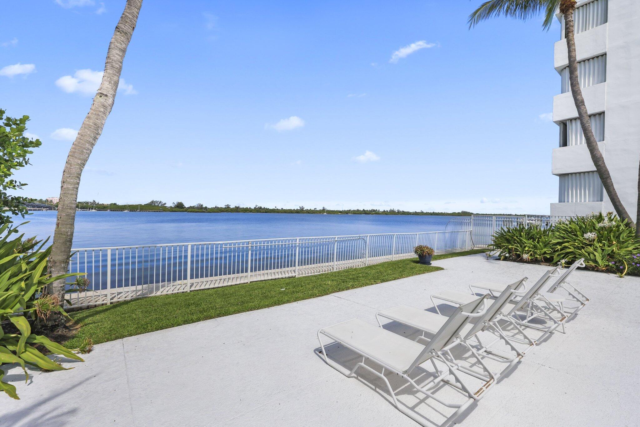 Waterfront patio with white chaise lounges, tropical landscaping, and calm blue water beyond the railing.