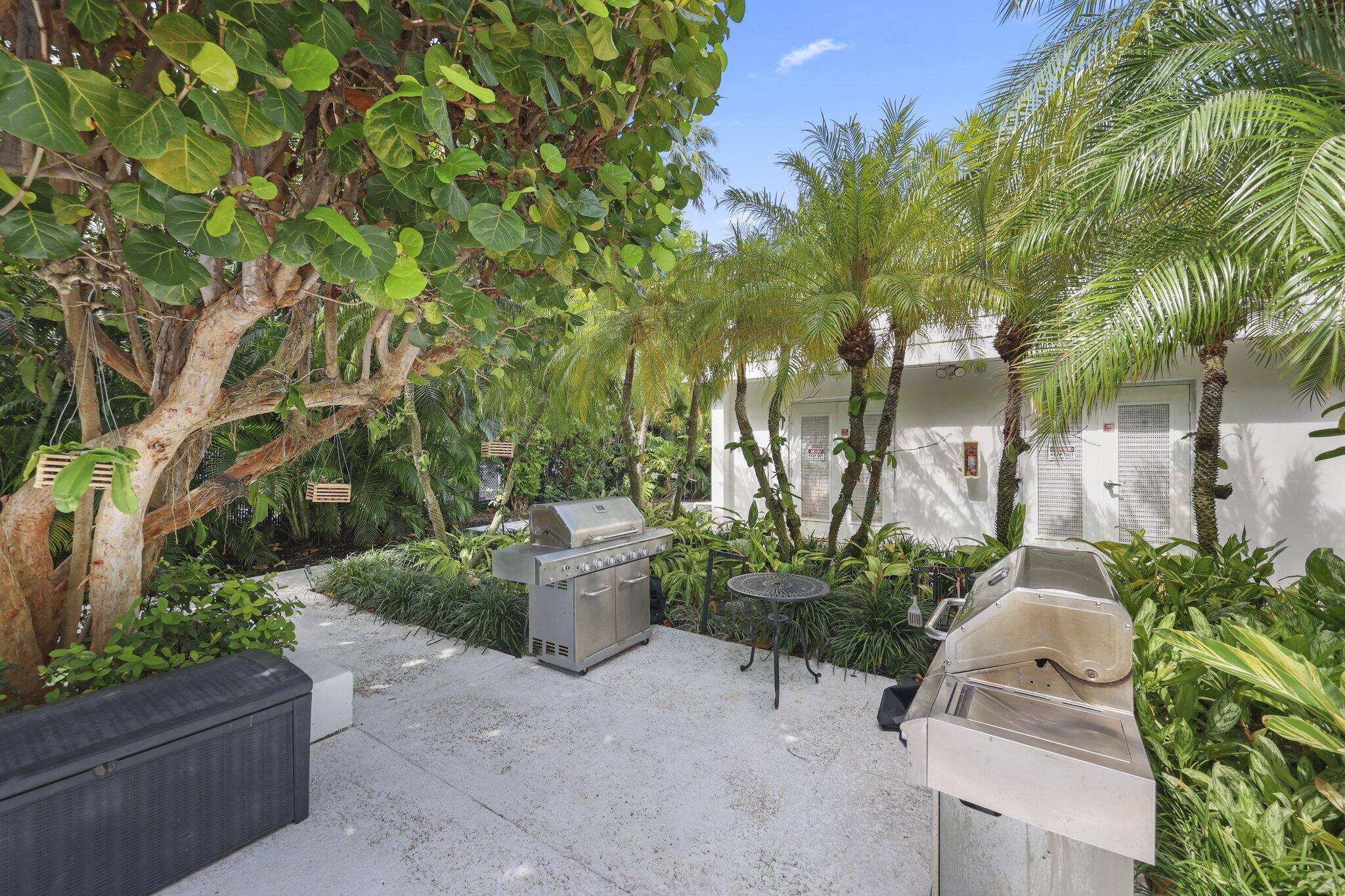 Outdoor patio with two stainless steel grills, a small table, and lush tropical landscaping.