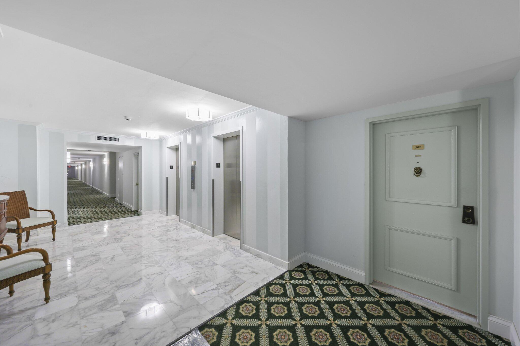 Condominium hallway with unit 302’s paneled door, elevators, marble floor, and patterned green carpet.