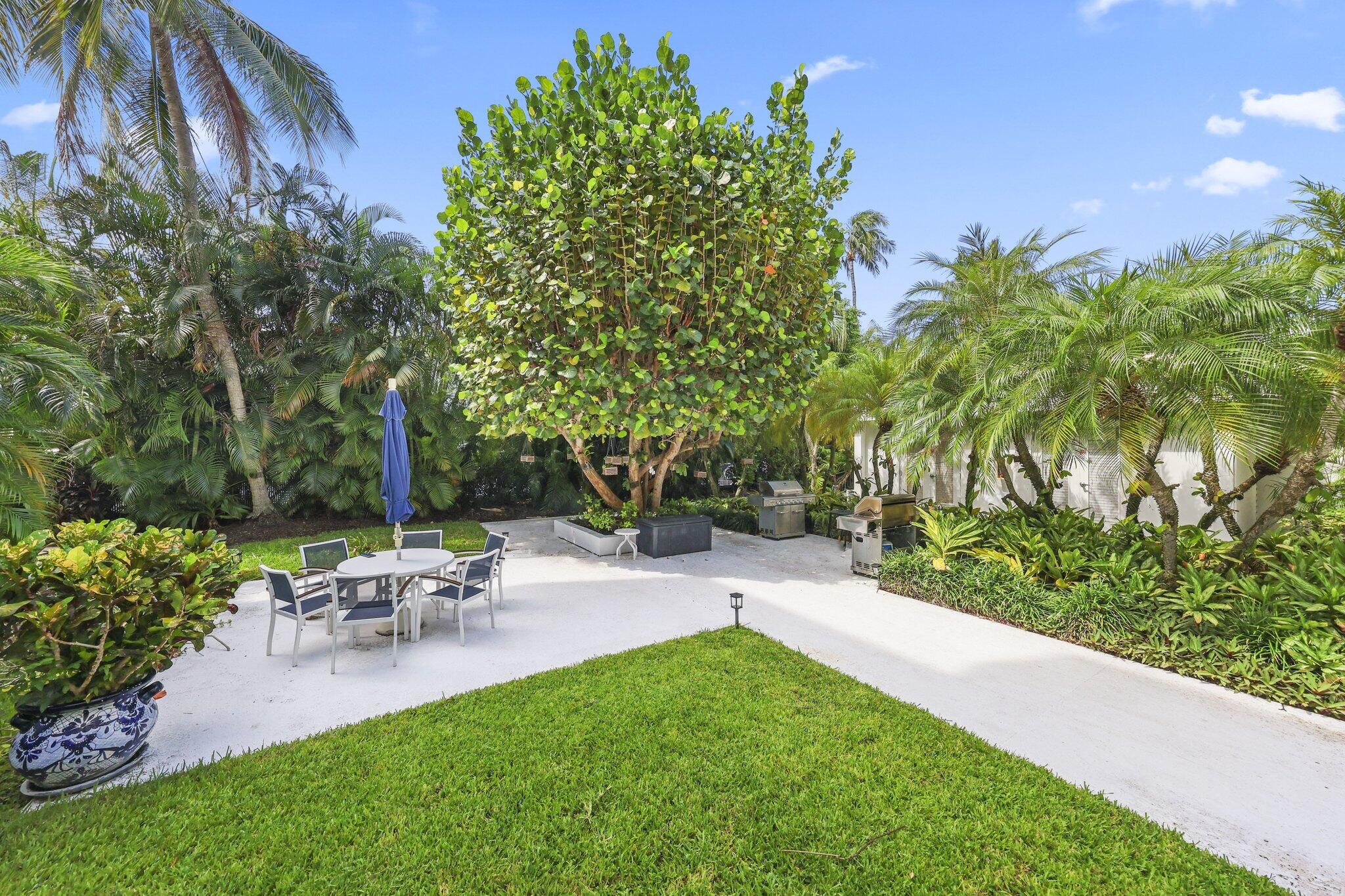Tropical garden patio with outdoor dining set, closed umbrella, and grills beside a lawn.