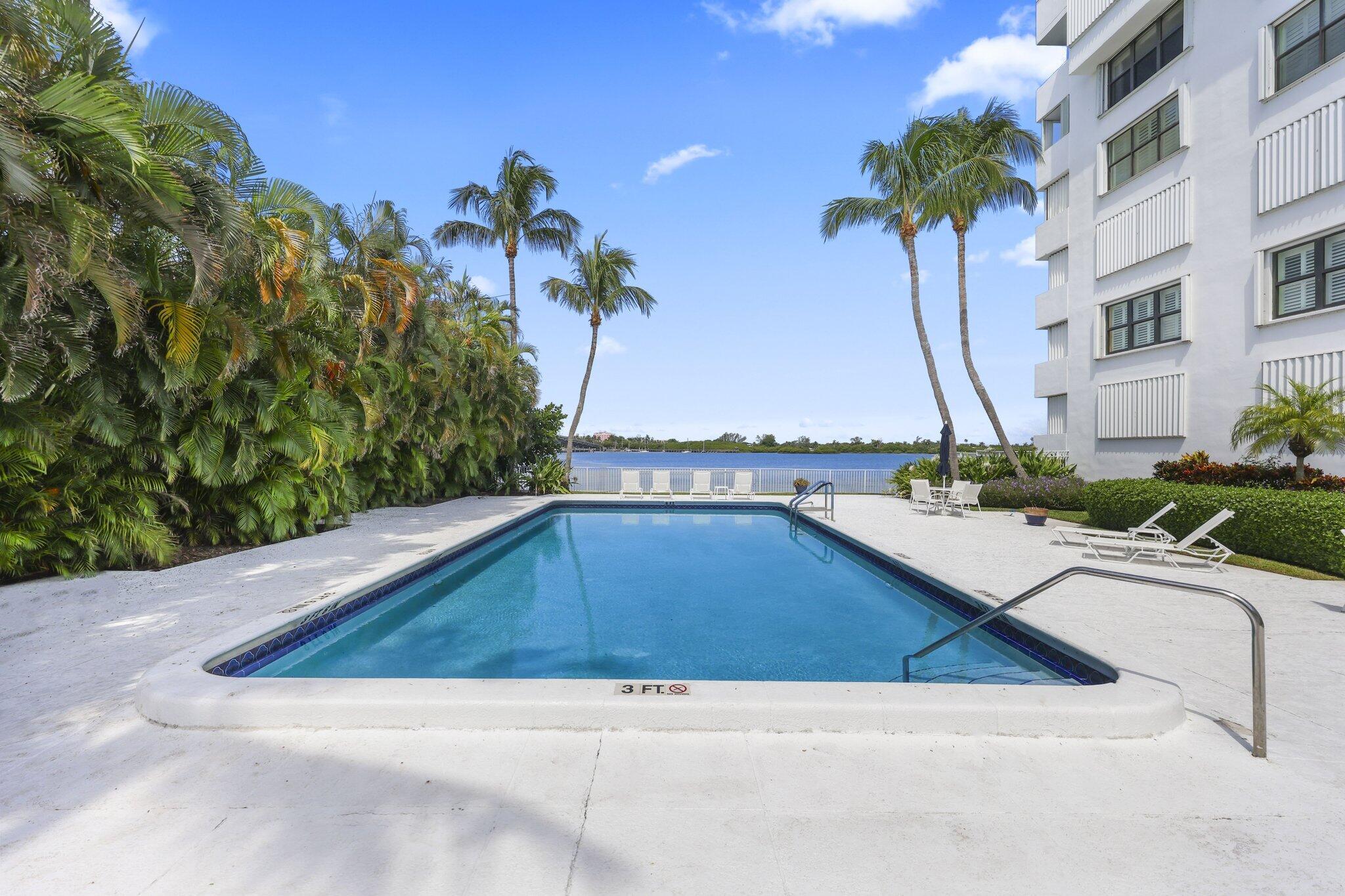 Condominium pool bordered by palm trees, with lounge chairs and a waterfront view.